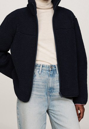 Persoon draagt een zwart fleecejack over een beige coltrui, ingestopt in lichtblauwe hoge taillejeans, staand tegen een effen achtergrond.