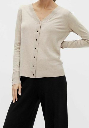 Femme portant un cardigan beige à col en V avec des boutons et des manches longues, associée à un pantalon large noir, posant avec une main sur la hanche.