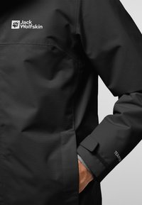Chaqueta negra impermeable de Jack Wolfskin con logo en el pecho y puños ajustables de Velcro en la manga, con la mano en el bolsillo.