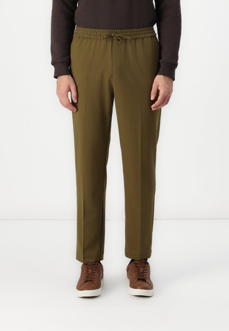 Olivgrüne tapered Hose mit elastischem Bund und Zugbanddetails. Glattes Material mit leichtem Glanz, kombiniert mit braunen Turnschuhen.
