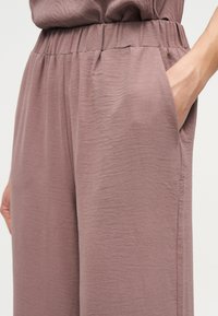 Μωβ υφασμάτινο παντελόνι με ελαστική μέση, με πλαϊνή τσέπη όπου ένα χέρι εισέρχεται μερικώς.