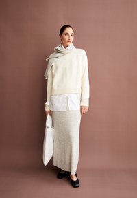 Crèmekleurige trui, witte blouse, grijze met patroon geslaagde rok en zwarte klompen. Accessoire: grijze sjaal. Houdt een witte tote bag vast. Bruine achtergrond.