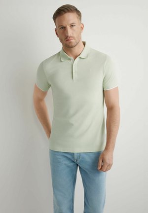 Mężczyzna stojący przy białej ścianie, ubrany w jasnozieloną koszulkę polo z krótkim rękawem i jasnoniebieskie dżinsy, patrzący przed siebie z neutralnym wyrazem twarzy.