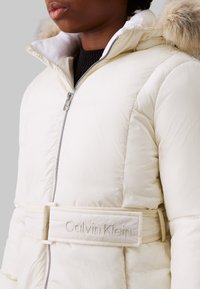 Giubbotto invernale color crema con zip frontale, cappuccio foderato in pelliccia e vita con cintura, caratterizzato da logo ricamato "Calvin Klein". Tessuto liscio.