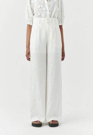 Kvinde iført høj-taljede, vidde hvide bukser og en gennemsigtig hvid bluse med blomstrede påsyede detaljer, stående mod en ensfarvet baggrund.