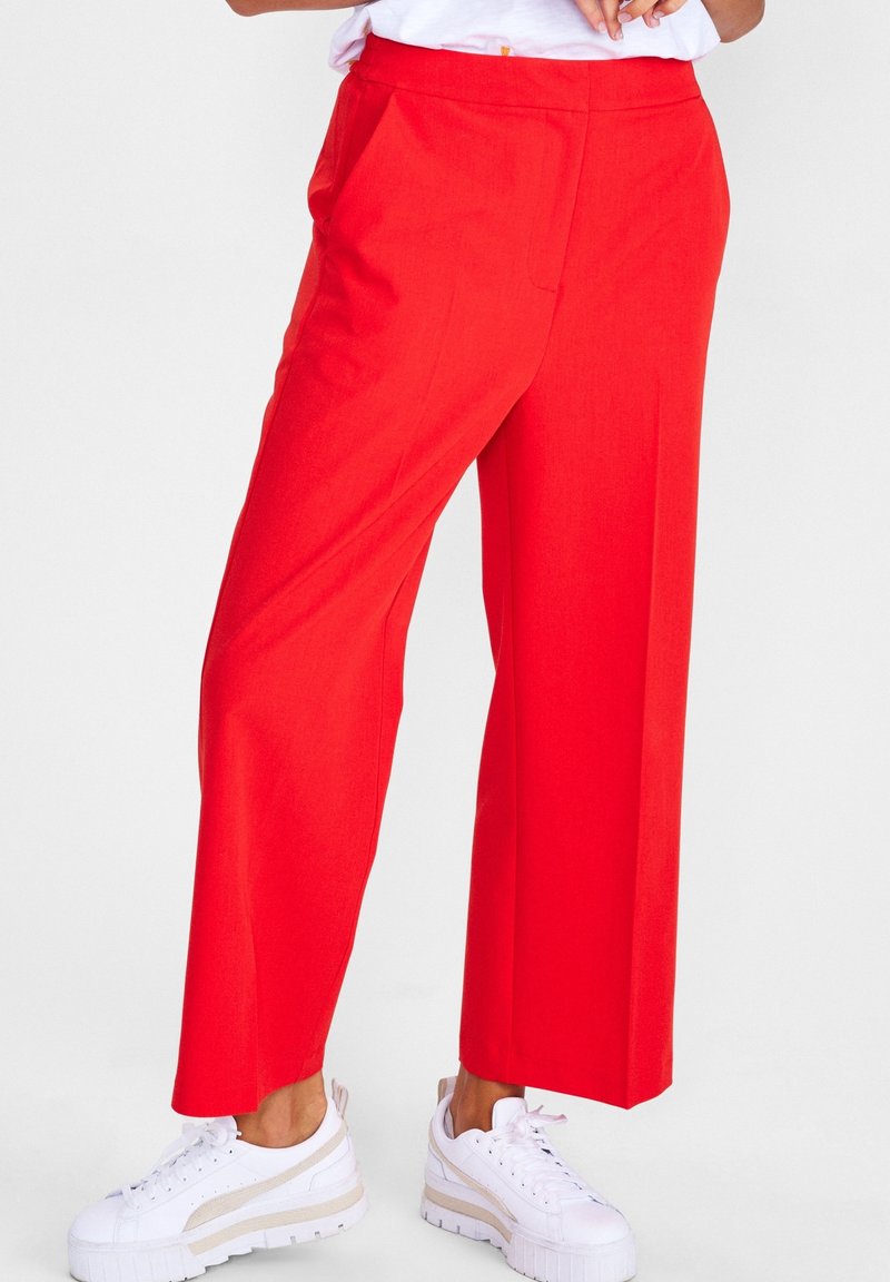 Personne portant un pantalon large rouge vif et des baskets blanches, debout devant un fond clair uni.