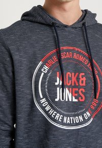 Felpa a righe blu navy con cappuccio e cordini. Presenta un logo centrale con testo rosso e bianco, inclusi "JACK & JONES" e ulteriori dettagli testuali circolari.