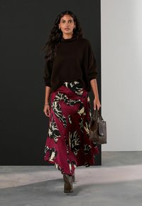 Bruine oversized trui, bordeauxrode bloemenrok met zwarte en crèmekleurige patronen, bruine handtas en bruin hoge laarzen op een eenvoudige achtergrond.