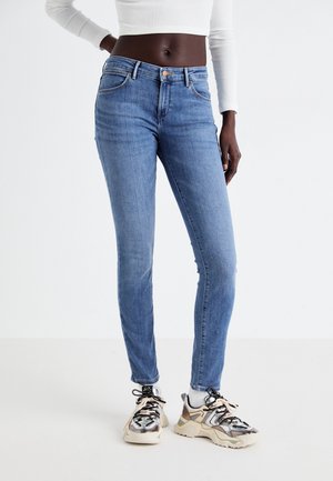 Jean skinny bleu présentant un style classique à cinq poches, délavé léger et détails de couture subtils, associés à des sneakers massives.