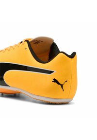 Botín de fútbol sintético amarillo con acentos de goma negra, que presenta un diseño aerodinámico, rayas en contraste y un logo negro de Puma en el talón.
