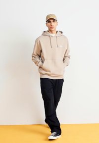 Hanorac bej cu buzunar frontal, glugă cu șnur și logo mic. Pantaloni cargo negri și adidași negri cu accente albe. Fundal galben.