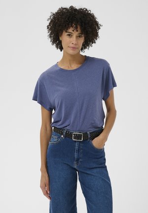 Femme aux cheveux bouclés portant une chemise bleue à manches courtes rentrée dans un jean taille haute bleu avec une ceinture tissée noire, main dans la poche.