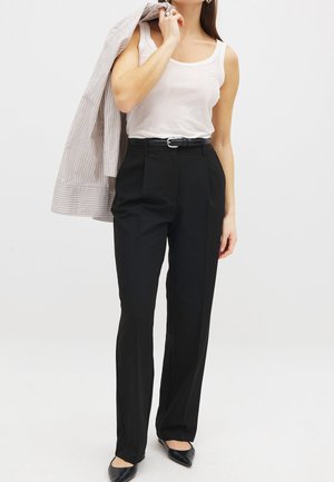 Kobieta w białym topie na ramiączkach, czarnych spodniach z wysokim stanem i paskiem, czarnych szpiczastych butach, trzymająca na ramieniu pasiastą marynarkę.