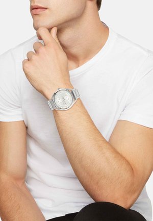 Silberner Edelstahluhren mit rundem Ziffernblatt, strukturiertem Zifferblatt und mehreren Zählerdials. Wird an einer Hand gegen ein einfaches weißes Hemd getragen.