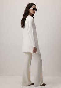 Sieviete ar garām tumšām matiem, tērpusies baltā garās piedurknes tunikā, krēmkrāsas platpirkstu biksēs, brūnās šaurās kurpēs un tumšās saulesbrillēs uz viendabīga fona.