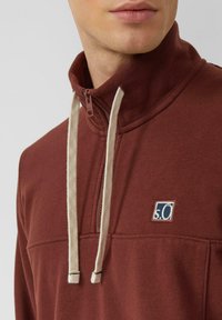 Maglione bordeaux con chiusura a mezza cerniera, dotato di cordoncini crema e un piccolo logo blu. Realizzato in un tessuto morbido e strutturato.