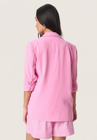 Rosa Blazer mit gekrempelten Ärmeln, der eine glatte Textur und eine taillierte Passform aufweist. Getragen über passenden gestreiften Shorts, von hinten gezeigt.
