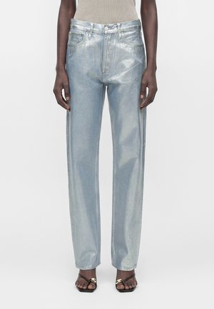 Modèle portant un jean bleu clair coupe ample avec un éclat métallique, accompagné d’un haut beige et de sandales noires ouvertes à l’avant avec un détail chaîne dorée.