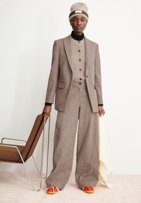 Brauner Wollanzug mit Textur, weiten Hosen und enger, geknöpfter Oberteil, kombiniert mit einem cremefarbenen Schal und bunten Sneakern. Modell hält einen Stuhl.