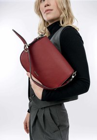 Red leather handbag with textured surface, curved design, silver hardware, and a detachable strap, held against a gray outfit.