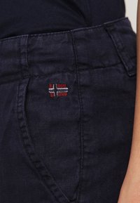 Detailní záběr na tmavě modrou kapsu kalhot s malým vyšitým červeným, bílým a černým křížem, připomínajícím nordickou vlajku, na horní straně.
