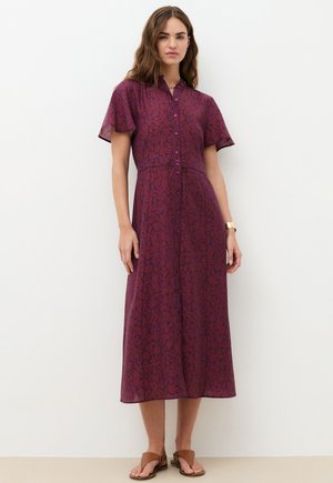 Donna che indossa un vestito midi viola con fantasia, bottoni sul davanti, maniche corte e sandali marroni, in piedi su uno sfondo bianco.