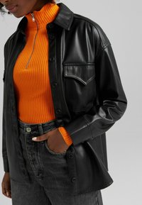 Černá kožená košile s kapsou na přední straně, oblečená přes jasně oranžový žebrovaný svetr. Šedé džíny s vysokým pasem doplňují celý outfit.