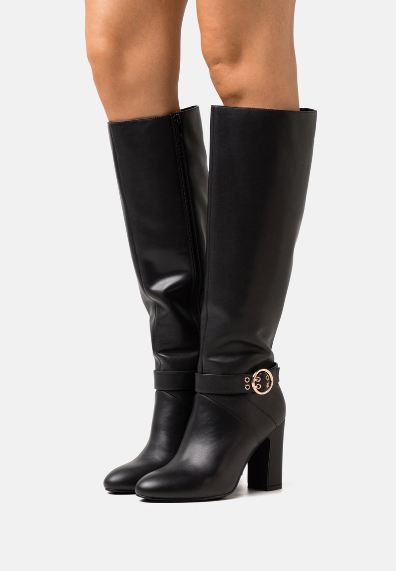 Bottes en cuir noires montantes jusqu'aux genoux avec talons carrés et détail boucle dorée, portées sur des jambes nues sur fond blanc.