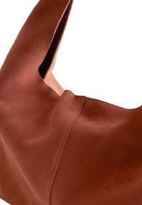 Braune Wildleder-Schultertasche mit weicher Textur, einzigartiger Form und minimalistischem Design. Verfügt über sichtbare Nähte und eine offene Oberseite.