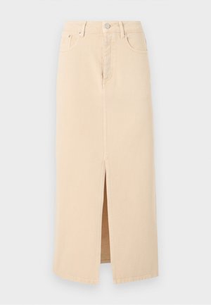 Kvinde iført beige jakke og nederdel med bølget kant, der holder en lille grøn håndtaske og står mod en ensfarvet lys baggrund.