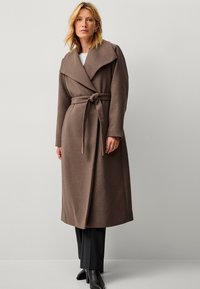 Cappotto lungo marrone realizzato in tessuto morbido, con un ampio colletto, vita regolabile e maniche lunghe. Abbinato a pantaloni neri e stivali.