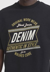Černé bavlněné tričko s výrazným bílým a zlatým textem. Design obsahuje akcenty "DENIM", "AUTENTICKÝ STYL" a "OŠETŘENÉ JÁDRO".
