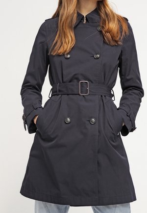 Vrouw die een zwarte dubbelrijige trenchcoat met riem draagt, handen in de zakken en lichtblauwe spijkerbroek, bij de nek en knieën bijgesneden.