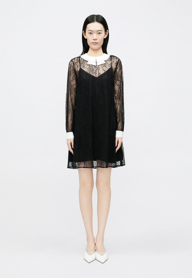 Svart kjole i blonder med lange ermer, V-utringning og hvit Peter Pan-krage. Teksturert mønster over hele kjolen og en vid silhuett med knelengde.