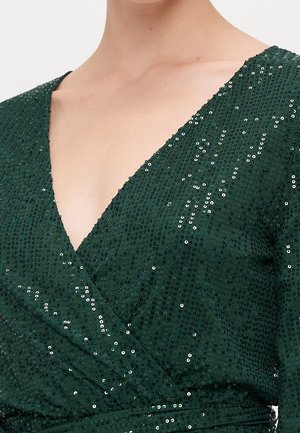 Primer plano de una persona que lleva un vestido de lentejuelas verde oscuro con escote en V pronunciado, estilo cruzado en la parte delantera y mangas largas.