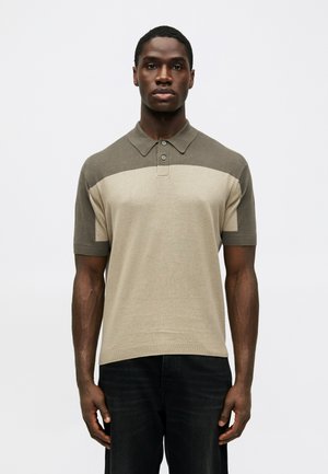 Uomo che indossa una polo a maniche corte bicolore beige e verde oliva abbinata a pantaloni neri, in piedi davanti a uno sfondo bianco semplice.