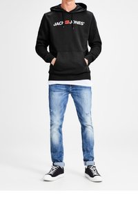 Man som bär en svart Jack & Jones hoodie med framficka, ljusblå uppvikt jeans och svarta sneakers med vita sulor och snören.