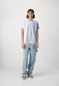T-shirt alb cu mânecă scurtă și logo mic, blugi albaștri deschiși, adidași din suede gri și o șapcă neagră, stând în fața unui fundal simplu.