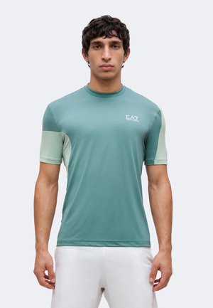 Bărbat purtând un tricou sport cu mâneci scurte, în două nuanțe – teal și verde deschis, cu logo-ul EA7, stând cu fața către înainte, pe un fundal simplu și deschis la culoare.