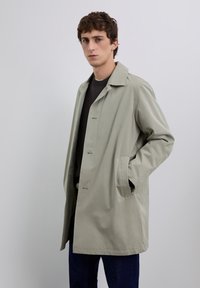 Jeune homme portant un trench-coat beige clair par-dessus un pull sombre et un pantalon foncé, se tenant avec une main dans la poche de son manteau devant un fond uni.