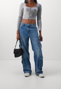 Maglione grigio corto con zip, jeans cargo blu con tasche laterali, sneakers bianche e una piccola borsa nera a spalla.