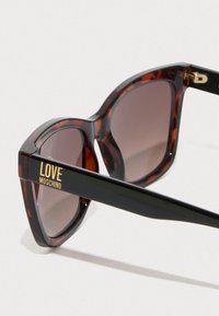 Kilpkonnakilbi päikeseprillid, mille must külgehaaraja on kaunistatud kuldsete "LOVE MOSCHINO" tähtedega ja tumedate objektiividega. Nurgeline kuju ja sile tekstuur.