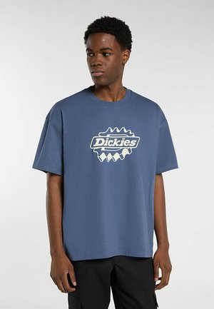 Joven con una camiseta azul holgada de Dickies y pantalones negros, de pie frente a un fondo claro y simple, mirando de lado.