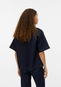 Mörkblå denimskjorta med korta ärmar, avslappnad design, synliga sömdetaljer och en lös passform, sedd bakifrån.