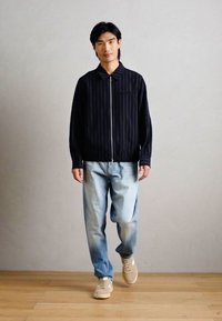 Παλτό ναυτικού στυλ με ρίγες, ελαφρύ ύφασμα, κλείσιμο με φερμουάρ, γιακάς; φορεμένο με ανοιχτό γαλάζιο τζιν ξεθωριασμένο και μπεζ αθλητικά παπούτσια σε ανοιχτό ξύλινο δάπεδο.