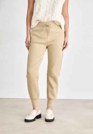 Beige Jogginghosen mit elastischen Bündchen und Kordelzugbund, kombiniert mit cremefarbenen Slippern mit schwarzer Plateausohle und abgerundeter Spitze.