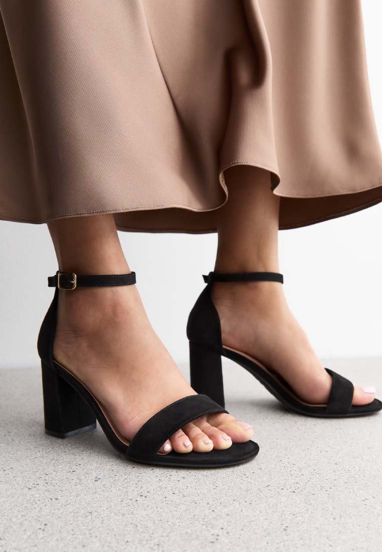Sandalias de tacón alto de ante negro con un grueso tacón cuadrado, correa en el tobillo y diseño de punta abierta, sobre un suelo texturizado claro.