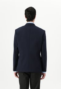 Blazer azul marino con un diseño estructurado, que presenta una abertura trasera sencilla y detalles de botones en las mangas, llevado sobre una camisa azul clara.
