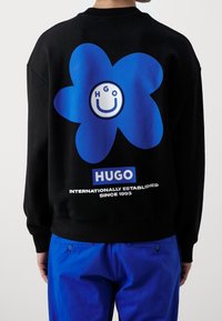 Černá mikina s velkým modrým květinovým motivem a usmívající se tváří s bílým nápisem "HUGO" nad textem "MEZINÁRODNĚ ZALOŽENO OD ROKU 1993."