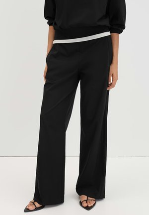 Femme portant un pantalon noir à jambes larges, les mains dans les poches, et des sandales noires à brides avec des bouts pointus sur fond uni.